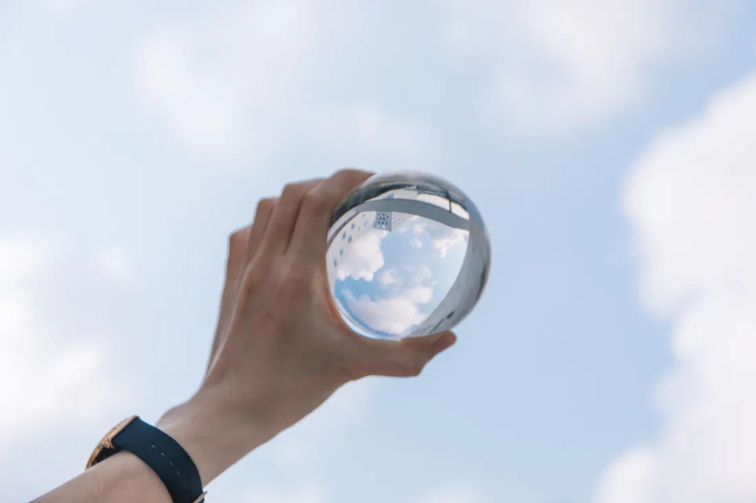 Hand hält eine Glaskugel vor bewölktem Himmel, darin spiegelt sich ein Gebäude